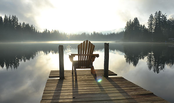 Esta escena bucólica de una silla vacía sobre un muelle de madera sobre aguas tranquillas que reflejan árboles y nubes ilustra el tema Los poderes de la paz, en editoriallapaz.