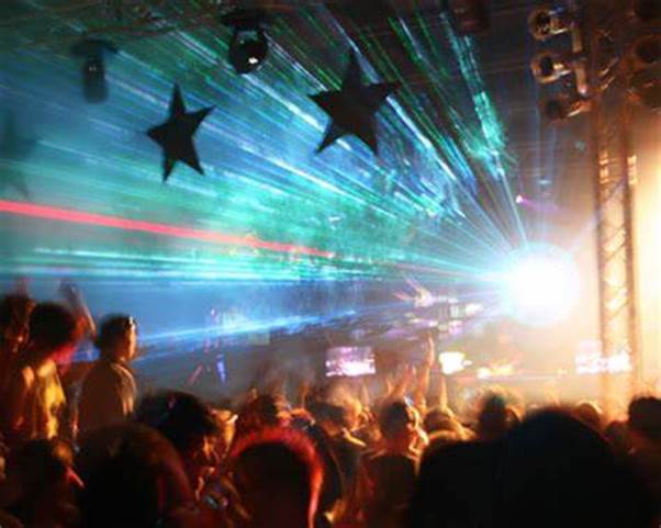 Una imagen de una muchedumbre de personas de espalda observando un espectáculo de entretenimiento, proyectándose de una luz blanca intensa en la tarima rayos verde blancos, con estrellas negras flotando en la parte superior del cradro.