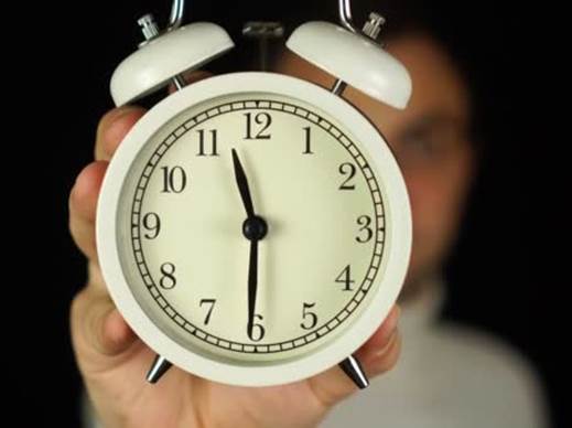 Fotografía de un reloj despertador, sostenido en la mano derecha de un varón, cuyas manecillas marcan las 11:30 p.m., teniendo el reloj dos cláxones y una cara blanca con números negros.