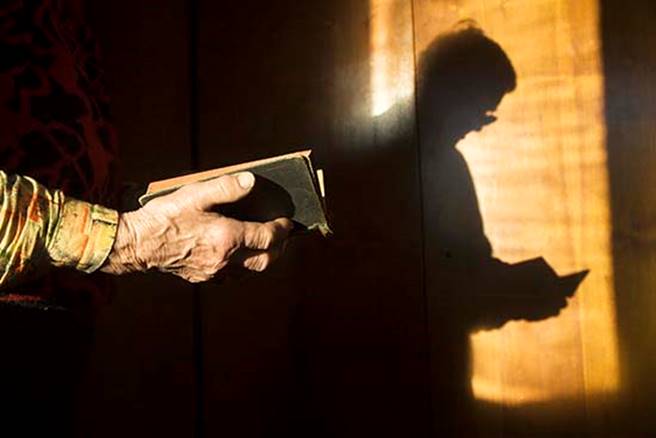 Fotografía de manos varoniles que sostiene una Biblia abierta, la sombra de ambos proyectada en una ventana de colores pardos suaves.