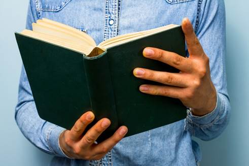 Fotografía de cerca de manos de un varón vestido de camisa azul de manga larga que sostienen un Biblia abierta.
