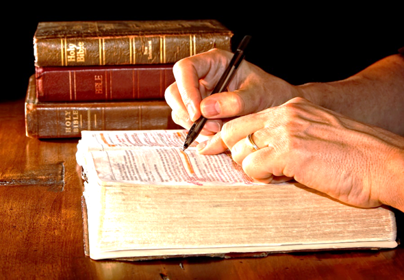 Foto de manos sobre una Biblia abierta con otros libros sobre un escritorio.