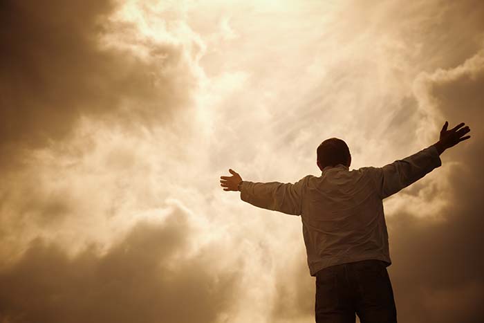 Esta gráfica de un varón a espaldas con los brazos levantados ante un espacio con una luz blanca aumentándose en intensidad entre nubes ilustra el tema Vivo en la tierra al aparecer Cristo por Segunda Vez, en editoriallapaz.org.
