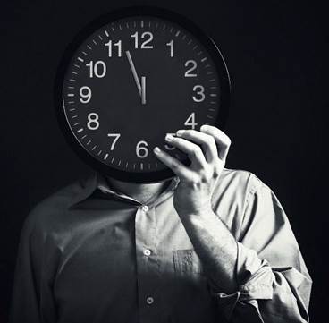 Una fotografía de un varón que sostiene un reloj grande negro con números blancos frente a su cara, marcando las manecillas casi, casi la medianoche.