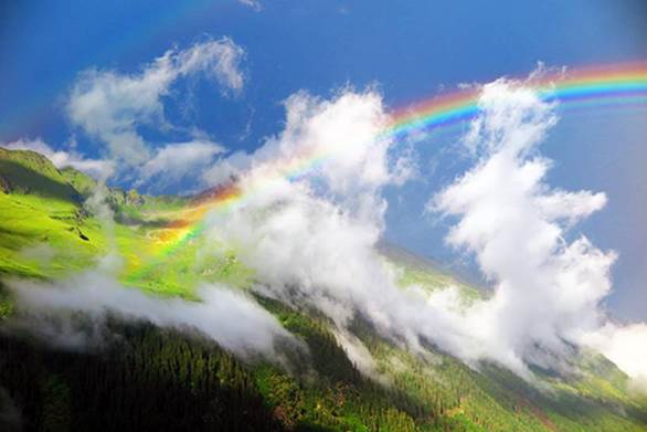 Fotografía de un arco iris entre pequeñas nubes blancas sobre la ladera de una montaña.