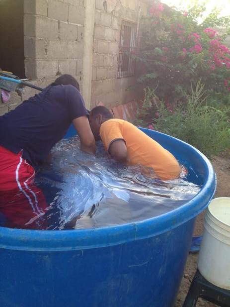 Fotografía de dos varones en el acto de bautizar por inmersión en agua a una persona en una gran bañera azul.