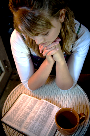 Fotografía tomada desde arriba de una mesita donde hay una Biblia abierta y una taza de café y sentada a la mesa una dama joven que lee la Biblia.