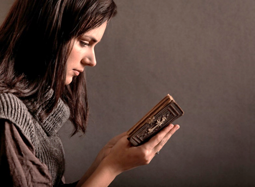 Fotografía de una dama joven que sostiene una Biblia abierta en las manos mientras la lee atentamente, contra un trasfondo pardo oscuro.