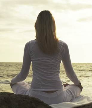 Una dama sentada frente al mar, con sus piernas cruzadas, practida la Yoga.