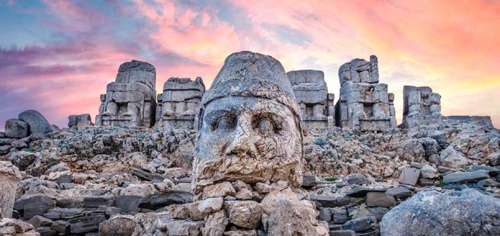 Una fotografía de las ruinas del Monte Nimrod en Turquía con una enorme cabeza en piedra en primera plana y detrás de ella porciones de enormes estatuas contra un trasfondo de nubes rosadas y cielo azul.