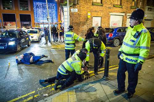 Fotografía tomada de celebrantes del Nuevo Año 2018 en Manchester, Inglaterra, con tres policiás que detieene a un sujeto y un varón tirado en la calle con una botella cerveza cerda de él, gráfica para el tema Cómo los británicos abordan la fe hoy día: de indiferencia a hostilidad abierta, en editoriallapaz.org.