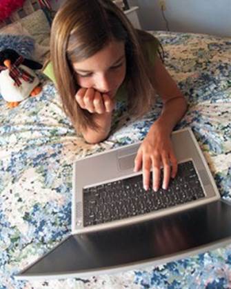 Fotografía de una niñita echada sobre su cama con una computadora laptop que ella va usando sin supervisión.